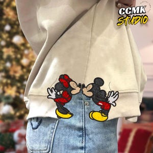 May include: Beige sweatshirt with a Mickey and Minnie Mouse kissing patch on the back. The patch features the iconic characters in black, red, and yellow. The text "CCMK Studio" is visible in the upper right corner.