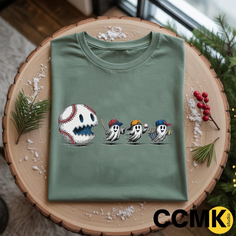May include: Sage green t-shirt featuring a baseball-themed design. The graphic includes a baseball with a ghost face, followed by three ghost figures wearing baseball caps and holding a bat, a baseball, and popcorn. The shirt is folded on a wooden surface.