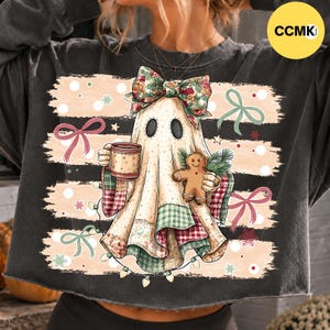 May include: A charcoal gray sweatshirt featuring a graphic of a ghost wearing a plaid bow, holding a mug and a gingerbread cookie. The design includes horizontal brush strokes, green and red bows, and string lights against a light background.