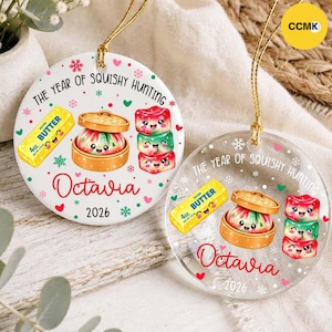 Könnte beinhalten: Zwei runde Ornamente, eines aus weißer Keramik und eines aus klarem Acryl, mit dem Text THE YEAR OF SQUISHY HUNTING, Octavia und 2026. Die Grafiken zeigen ein gelbes 113g Butterstück, einen Knödel im Bambusdämpfer und drei gestapelte Spielzeuge.