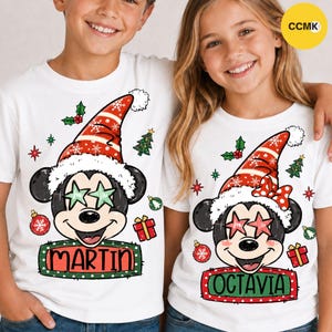 Peut inclure: Deux t-shirts blancs avec un motif de Mickey Mouse portant un bonnet de Père Noël rouge et blanc. Le t-shirt de gauche affiche le nom MARTIN, celui de droite le nom OCTAVIA. Les motifs incluent des lunettes en forme d'étoile, des sapins, des boules et des cadeaux.