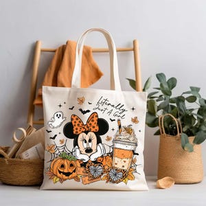 Pu&ograve; includere: Una borsa in tela color crema con una stampa grafica di un topo dei cartoni animati con un fiocco a pois, una zucca, una ragnatela e una tazza di caff&egrave;. Il testo "Literally Just A Girl" &egrave; stampato in nero sopra l'illustrazione.