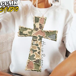 Può includere: Felpa bianca con una grande applicazione a forma di croce strutturata con un motivo mimetico nei toni del verde, marrone e rosa. Le parole "He is Risen" sono scritte verticalmente sulla croce. L'angolo in alto a sinistra ha il testo "CCMK STUDIO" in giallo e nero.