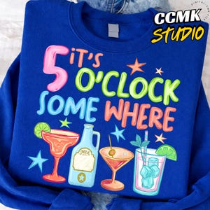 May include: A royal blue sweatshirt featuring the text "It's 5 O'Clock Somewhere" in vibrant colours. The design includes cocktail illustrations, a milk bottle, and stars. This is a casual wear item.
