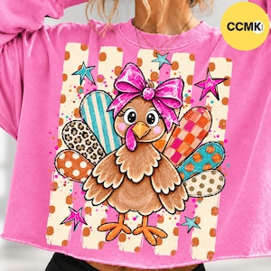 May include: A bright pink sweatshirt featuring a graphic of a brown turkey with a large pink bow on its head. The turkey has colorful patterned tail feathers including leopard print, stripes, and pumpkins. The background has vertical stripes.