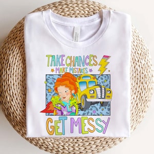 May include: A white t-shirt featuring a colorful graphic of a person with orange hair, a small green creature, and a yellow school bus. The text reads "TAKE CHANCES MAKE MISTAKES GET MESSY" with a yellow lightning bolt and small stars.