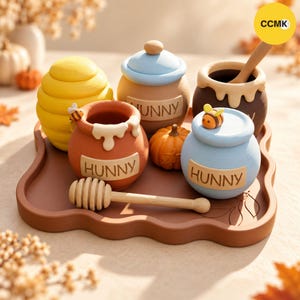 Puede incluir: Un conjunto de tarros de miel decorativos sobre una bandeja marrón, que incluye un tarro amarillo con forma de colmena, un tarro marrón con un cazo de madera y dos tarros etiquetados como HUNNY en azul y naranja. Incluye una calabaza y abejas.