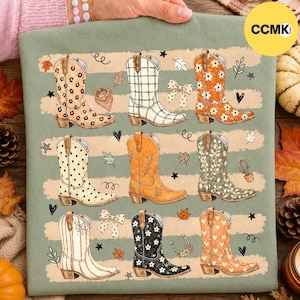 Puede incluir: Una camiseta verde salvia con un estampado gráfico de nueve botas vaqueras diferentes dispuestas en tres filas. Las botas muestran varios patrones, incluyendo estampado de leopardo, cuadros, lunares, motivos florales y rayas, intercalados con pequeños iconos de hojas, estrellas y corazones.