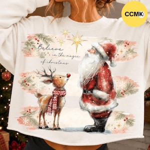 Op de afbeelding: Een crèmekleurig sweatshirt met een afbeelding van de Kerstman en een rendier. De tekst luidt "Believe in the magic of christmas" met een gouden ster erboven. Het ontwerp bevat aquarelachtige dennentakken, rode bessen en sneeuwvlokken.