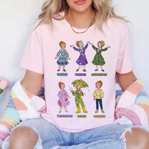 May include: A light pink short-sleeved t-shirt featuring a graphic print of a cartoon character in six different outfits labeled Monday through Weekend. The character wears various dresses, a raincoat with an umbrella, and casual clothing in colorful patterns.