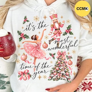 Puede incluir: Una sudadera con capucha blanca con un gráfico festivo que muestra una bailarina rosa, un cascanueces, un árbol de Navidad decorado, acebo y adornos. El texto, it's the most wonderful time of the year, está escrito en letra cursiva negra.