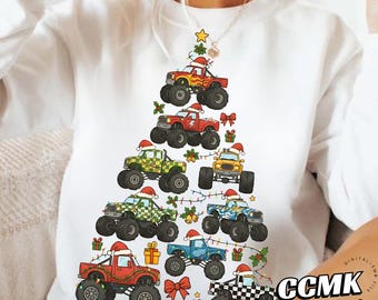 May include: White sweatshirt featuring a Christmas tree design made of monster trucks. Each truck is decorated with Santa hats, bows, and garland. The design includes gift boxes and a star on top. The text "CCMK STUDIO" is visible.