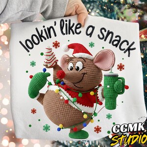 Op de afbeelding: Een wit kersthanddoek met een gehaakte muis met een kerstmuts, die een kerstboom en een groene mok vasthoudt. De tekst "lookin' like a snack" staat boven de muis, met sneeuwvlokken en de tekst "CCMK STUDIO" eronder.