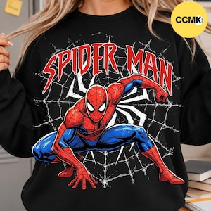 May include: A black crewneck sweatshirt featuring a graphic of the character Spider-Man in a crouching pose. The design includes a white spiderweb background and the text SPIDER MAN in bold red letters across the top of the chest area.