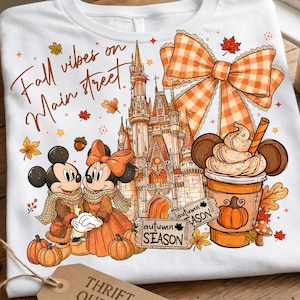 Puede incluir: Una camiseta blanca con un estampado gr&aacute;fico que incluye el texto Fall vibes on Main street, un castillo, dos ratones de dibujos animados con ropa de oto&ntilde;o, un lazo grande a cuadros naranja, una bebida de calabaza y dos tickets autumn SEASON.