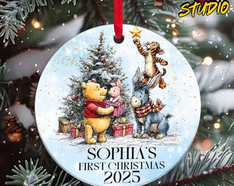 May include: A round Christmas ornament featuring Winnie the Pooh, Piglet, Eeyore, and Tigger around a Christmas tree. The ornament has the text "SOPHIA'S FIRST CHRISTMAS 2025" and is hanging from a red ribbon.