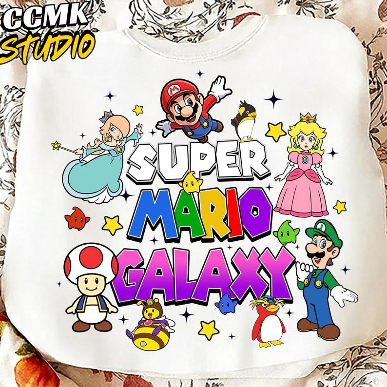 May include: White sweatshirt featuring a colorful Super Mario Galaxy graphic. The design includes Mario, Princess Peach, Luigi, and other characters. The text "CCMK STUDIO" is in the upper left corner.