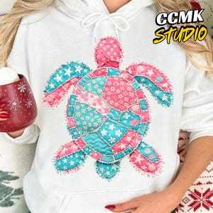 May include: White hoodie featuring a colorful patchwork turtle design. The turtle has pink, blue, and white patterned sections with star and floral accents. The hoodie has a drawstring and the text "CCMK STUDIO" in the upper right corner.