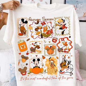 Puede incluir: Una camiseta blanca con un estampado gr&aacute;fico colorido de personajes de Disney en entornos oto&ntilde;ales. El dise&ntilde;o incluye a Mickey, Minnie, Donald, Daisy y Pluto con calabazas, hojas y bellotas. El texto dice Autumn Season y It's the most wonderful time of the year.