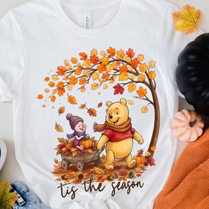 Puede incluir: Una camiseta blanca con un gr&aacute;fico de Winnie the Pooh y Piglet en una escena oto&ntilde;al. Pooh lleva una bufanda marr&oacute;n y una camisa roja, mientras Piglet est&aacute; sentado en un carro de madera lleno de hojas y una calabaza bajo un &aacute;rbol oto&ntilde;al.