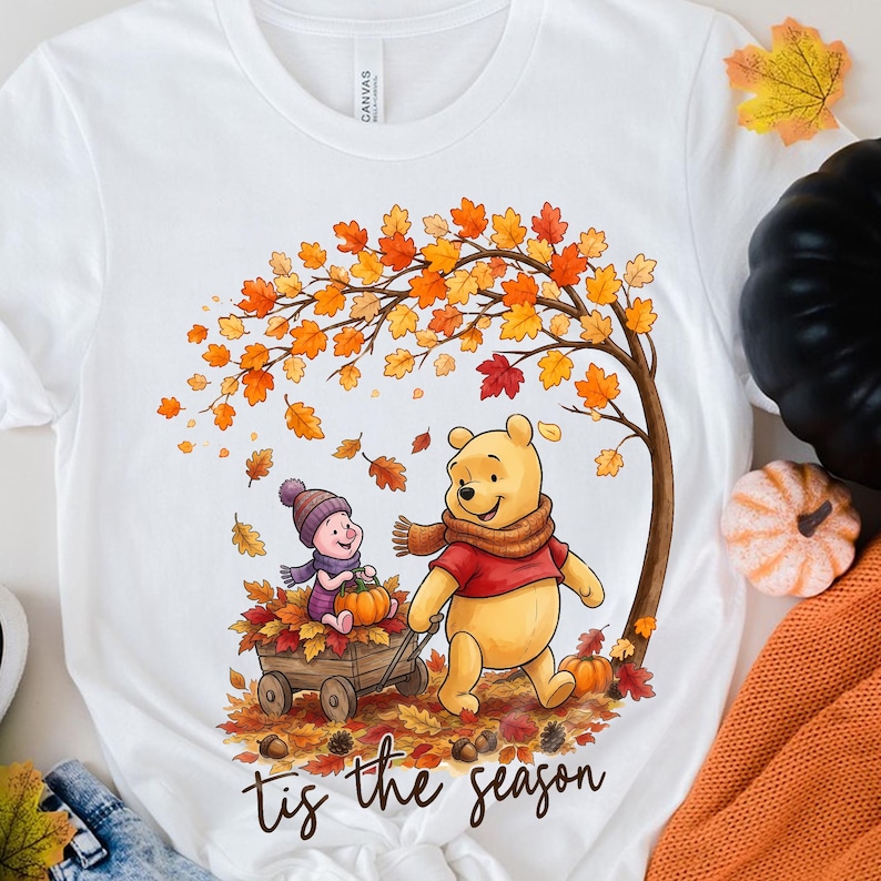 Puede incluir: Una camiseta blanca con un gr&aacute;fico de Winnie the Pooh y Piglet en una escena oto&ntilde;al. Pooh lleva una bufanda marr&oacute;n y una camisa roja, mientras Piglet est&aacute; sentado en un carro de madera lleno de hojas y una calabaza bajo un &aacute;rbol oto&ntilde;al.