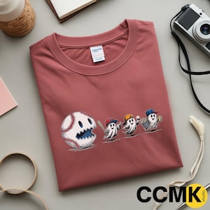 May include: A dusty rose-colored t-shirt featuring a baseball-themed graphic. The design includes a baseball with a smiling face and three ghost characters wearing baseball caps, holding a bat, a baseball, and popcorn.