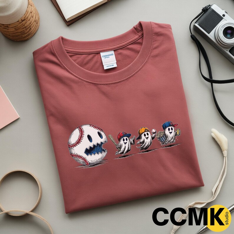 May include: A dusty rose-colored t-shirt featuring a baseball-themed graphic. The design includes a baseball with a smiling face and three ghost characters wearing baseball caps, holding a bat, a baseball, and popcorn.