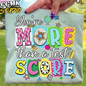 Op de afbeelding: Een lichtgroene sweatshirt met de tekst "You're More Than a Test Score" in een kleurrijk, speels ontwerp. De letters zijn in verschillende patronen, waaronder madeliefjes, sterren en luipaardprint. Schoolthema-afbeeldingen zijn verspreid.