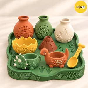 May include: A set of dinosaur-themed silicone kitchenware on a green tray, including three small vases, a cracked egg bowl, a volcano mold, a triceratops bowl, a dinosaur-shaped bowl, and a yellow spoon, all featuring embossed prehistoric motifs.