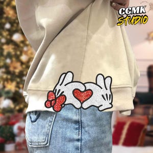 Puede incluir: Sudadera beige con un gráfico de dos manos enguantadas blancas formando un corazón con un lazo rojo a la izquierda. El texto "CCMK Studio" está en amarillo en la parte superior derecha.