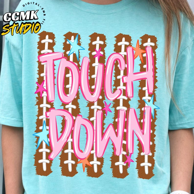 May include: A light blue t-shirt featuring a graphic print of five vertical brown football-textured stripes with white laces. The text "TOUCH DOWN" is printed in large pink letters over the stripes, accented by small light blue, pink, and orange stars.