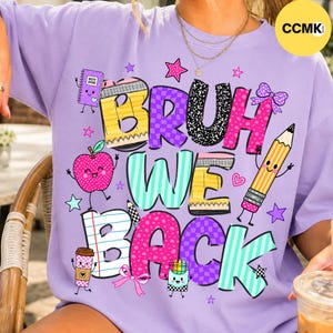May include: A light purple t-shirt featuring the text BRUH WE BACK in colorful, patterned block letters. The design includes cartoon illustrations of a notebook, a pink apple, a yellow pencil, a coffee cup, and a box of crayons, all with faces.