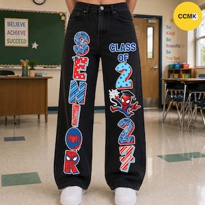 May include: Black denim jeans featuring a graphic print on the legs. The left leg displays the word SENIOR in red, white, and blue with spiderweb patterns. The right leg shows CLASS OF 2027 with a Spider-Man character and web designs.