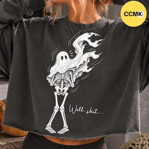 Könnte beinhalten: Ein anthrazitfarbenes, bauchfreies Sweatshirt mit einer weißen grafischen Illustration eines Skeletts, das in ein Geisterlaken gehüllt ist. Das Skelett hält die Hände vor der Brust und der Text "Well, shit..." steht in weißer Schrift darunter.