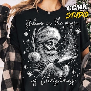 May include: Black t-shirt featuring a detailed illustration of Santa Claus with a long beard, glasses, and a fur-trimmed hat. The text "Believe in the magic of Christmas" is written in a script font, with snowflakes scattered around the image.