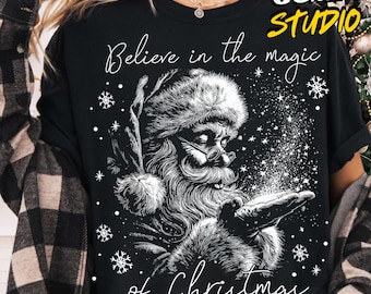 May include: Black t-shirt featuring a detailed illustration of Santa Claus with a long beard, glasses, and a fur-trimmed hat. The text "Believe in the magic of Christmas" is written in a script font, with snowflakes scattered around the image.