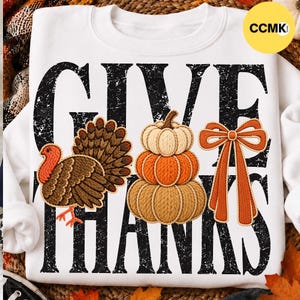 Op de afbeelding: Een wit sweatshirt met ronde hals en de zwarte, verweerde tekst GIVE THANKS. Het ontwerp bevat een bruine kalkoen, een stapel van drie pompoenen in beige en oranje tinten, en een oranje strik met een witte omlijning.