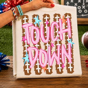 May include: A cream-colored t-shirt featuring a graphic print of brown football laces arranged in vertical stripes. The text TOUCH DOWN is printed in large, bubble-style pink letters, accented by small, scattered light blue and orange stars.