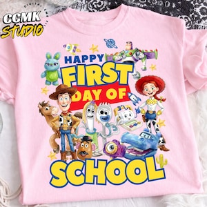 Può includere: Una t-shirt rosa chiaro con una grafica colorata dei personaggi di Toy Story, tra cui Woody, Jessie, Buzz Lightyear e Forky. Il testo Happy First Day Of School è scritto in grassetto blu e giallo con piccoli accenti a forma di stella.