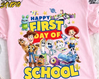 May include: A light pink t-shirt featuring a colorful graphic of Toy Story characters including Woody, Jessie, Buzz Lightyear, Forky, and others. The text reads Happy First Day Of School in bold blue and yellow letters with small star accents.