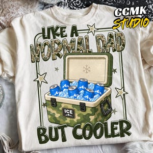 May include: Cream-colored t-shirt with the text "LIKE A NORMAL DAD BUT COOLER" in green and camouflage lettering. The graphic features a green camouflage cooler filled with blue cans and ice cubes. The shirt has a crew neck and short sleeves.