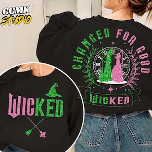 Può includere: Felpa nera con la scritta "WICKED" in glitter rosa e verde. Il design include un cappello da strega, una scopa e la frase "CHANGED FOR GOOD" con due figure di streghe. È presente anche un design a orologio.