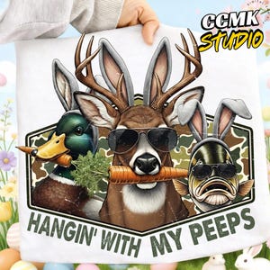 May include: White t-shirt featuring a humorous graphic of a duck, deer, and fish, each wearing sunglasses and bunny ears, holding carrots. The text "HANGIN' WITH MY PEEPS" is printed below the graphic. The CCMK STUDIO logo is in the upper right corner.