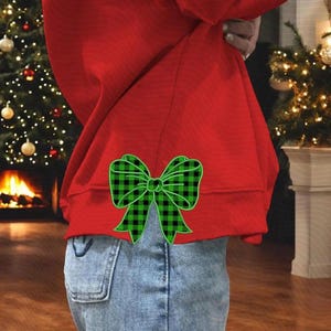 Könnte beinhalten: Ein rotes Sweatshirt mit einem grün-schwarz karierten Schleifendesign auf dem unteren Rücken, kombiniert mit einer blauen Jeans. Das Bild wurde in Innenräumen aufgenommen, mit einem Weihnachtsbaum und einem Kamin im Hintergrund.