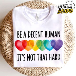 Op de afbeelding: Een wit t-shirt met de zwarte tekst BE A DECENT HUMAN IT'S NOT THAT HARD. Onder de eerste regel tekst staat een rij van zes overlappende harten in aquarel-stijl in rood, oranje, geel, groen, blauw en paars, die een regenboogverloop vormen.