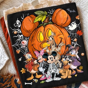 Puede incluir: Una camiseta negra con una calabaza naranja grande con orejas de Mickey Mouse, rodeada de telara&ntilde;as, murci&eacute;lagos y hojas de oto&ntilde;o. Debajo, personajes de dibujos animados aparecen con disfraces de Halloween como esqueleto y bruja.