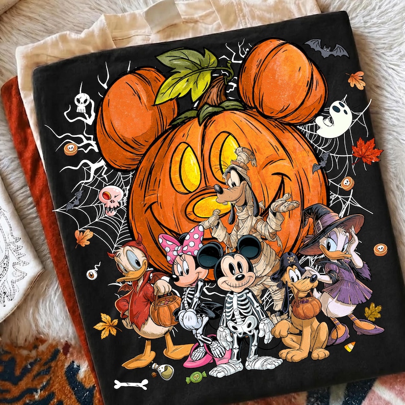 Puede incluir: Una camiseta negra con una calabaza naranja grande con orejas de Mickey Mouse, rodeada de telara&ntilde;as, murci&eacute;lagos y hojas de oto&ntilde;o. Debajo, personajes de dibujos animados aparecen con disfraces de Halloween como esqueleto y bruja.