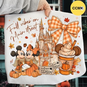 May include: A white t-shirt featuring a graphic print of a castle, two cartoon mice in autumn attire, a large orange plaid bow, and a cup of coffee. The text reads "Fall vibes on Main street" and "autumn SEASON" with pumpkins and leaves.