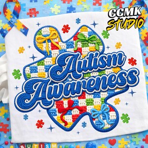 May include: White t-shirt with a blue, red, green, and yellow puzzle piece design. The words "Autism Awareness" are embroidered in blue. The shirt has a blue border and decorative bows. The image also includes the text "CCMK Studio".