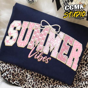 Puede incluir: Sudadera azul marino con las palabras "SUMMER Vibes" en rosa y blanco, adornada con perlas. Las letras tienen diseños de conchas marinas y flores. La sudadera está doblada sobre una tela con estampado de leopardo.